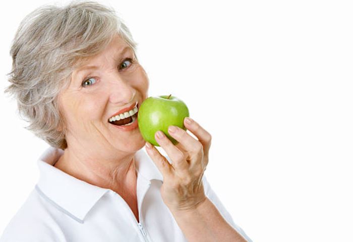 Elderly woman with dentures enjoying an apple, highlighting denture services in Newnan and Georgia.
